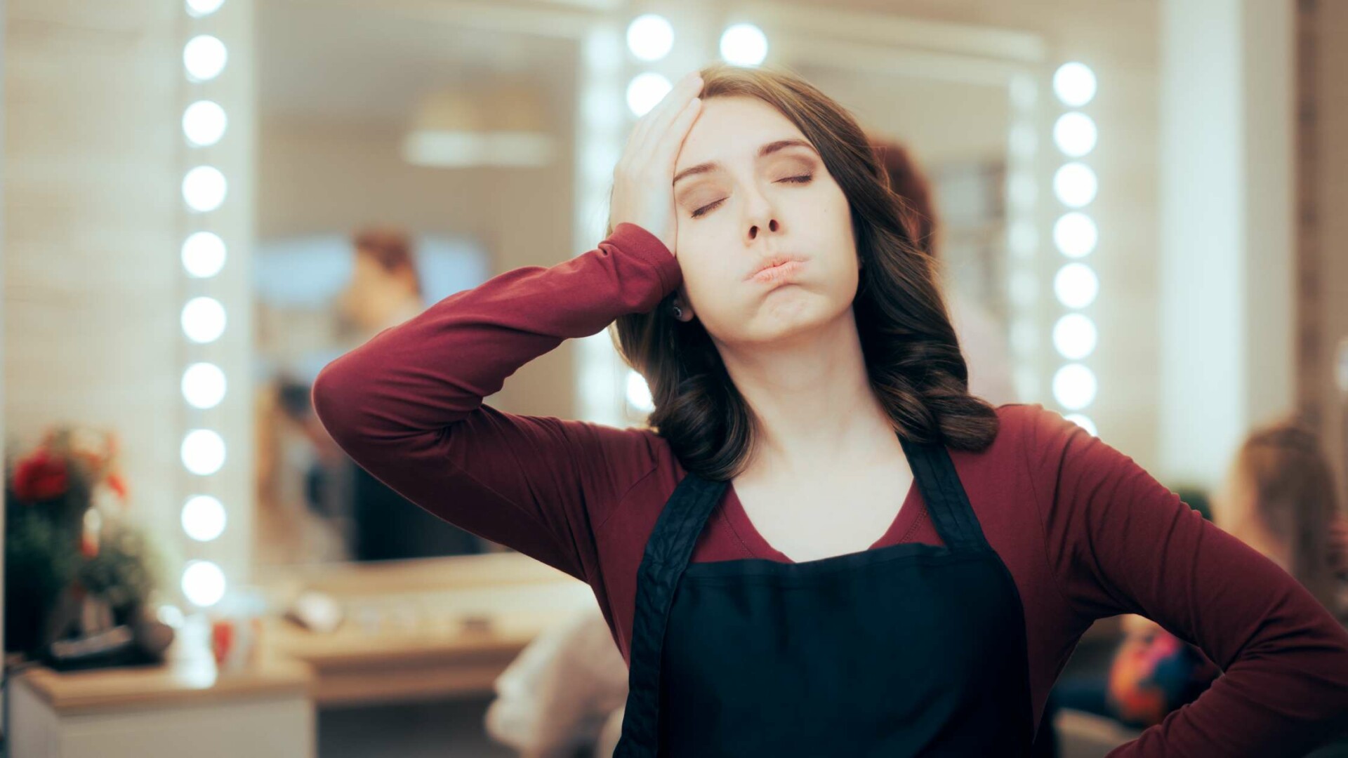 A stressed-out female hairdresser with one hand on her head and the other on her hip standing in front of a salon mirror.