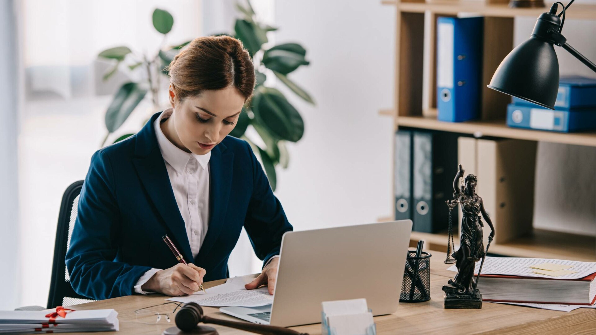 A female lawyer wearing a suit sits at a desk with a laptop while using a pen to write a note on a legal contract.