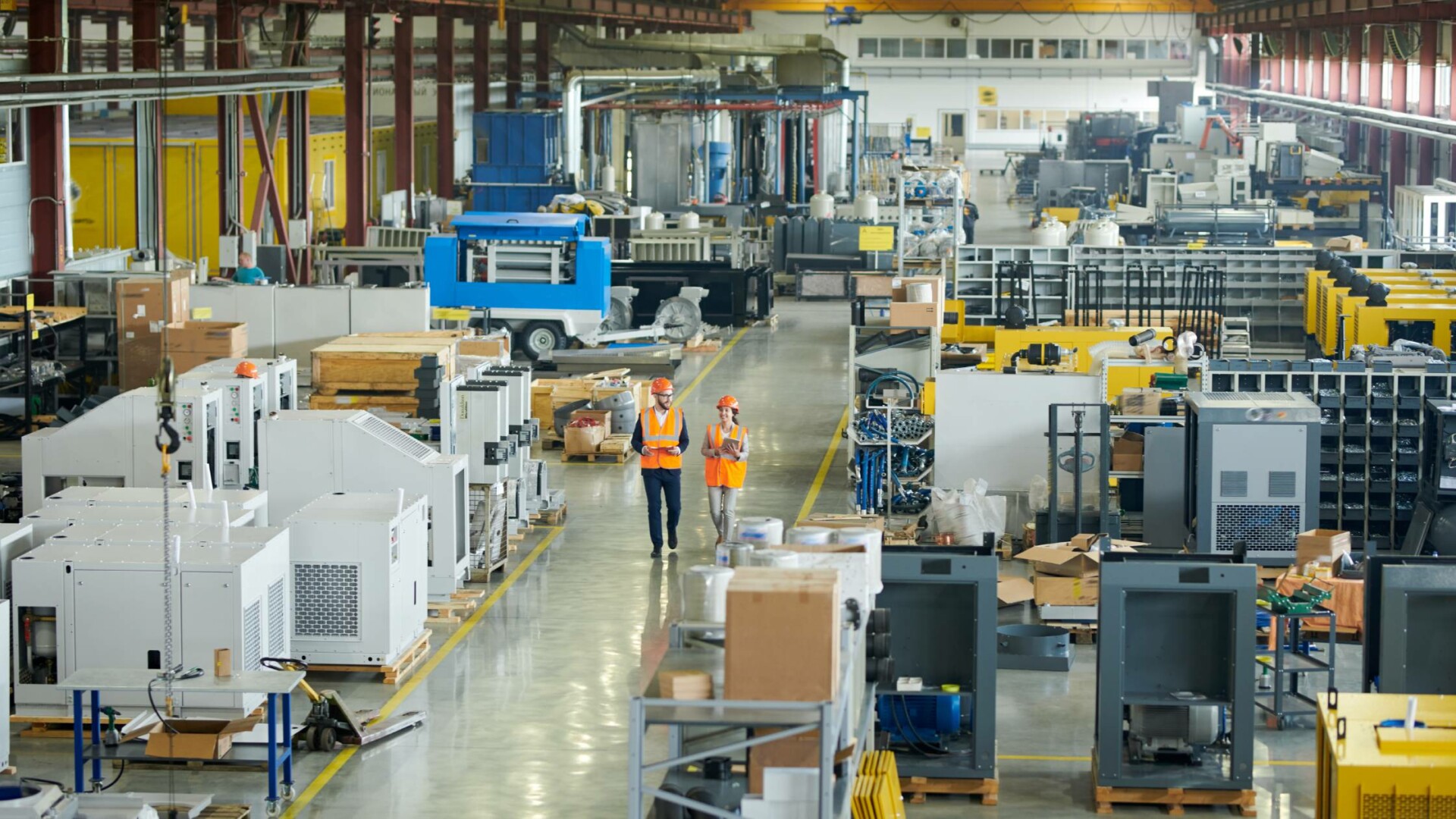 A bird's eye view of two employees walking through a manufacturing floor in orange safety vests and hard hats.