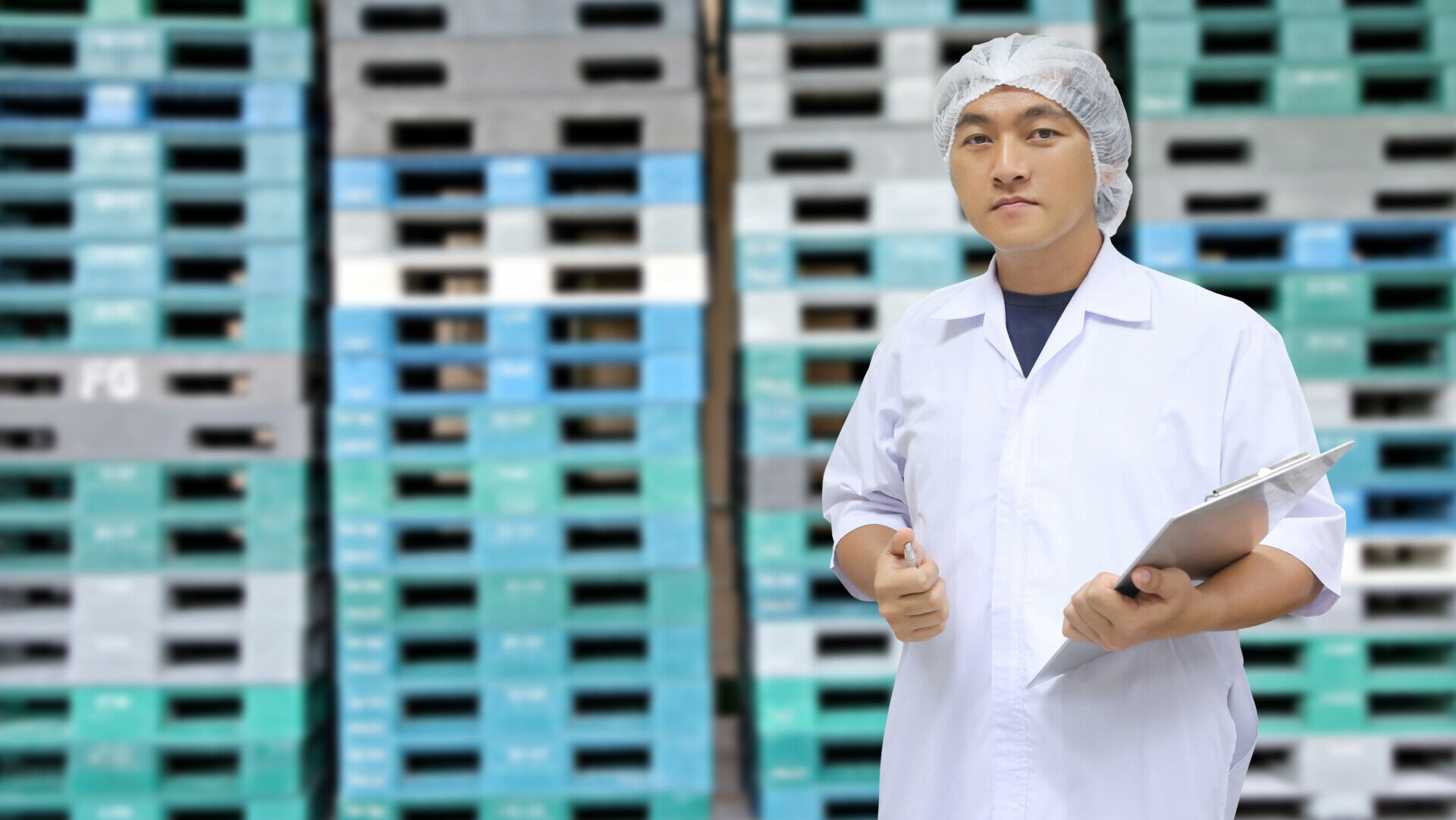 A man in a white coat and a hair net holding a clipboard and standing in front of several stacks of plastic pallets.