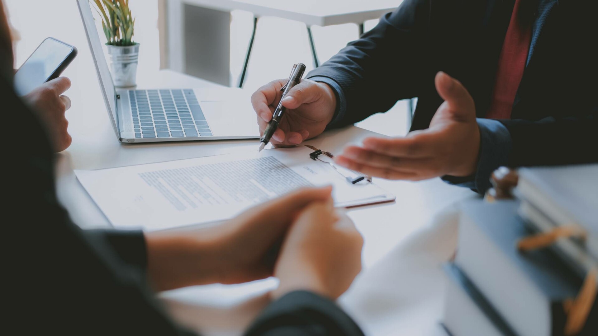 Three people sit at a desk with an open laptop, a document, and a stack of books. One person is holding a pen and talking.