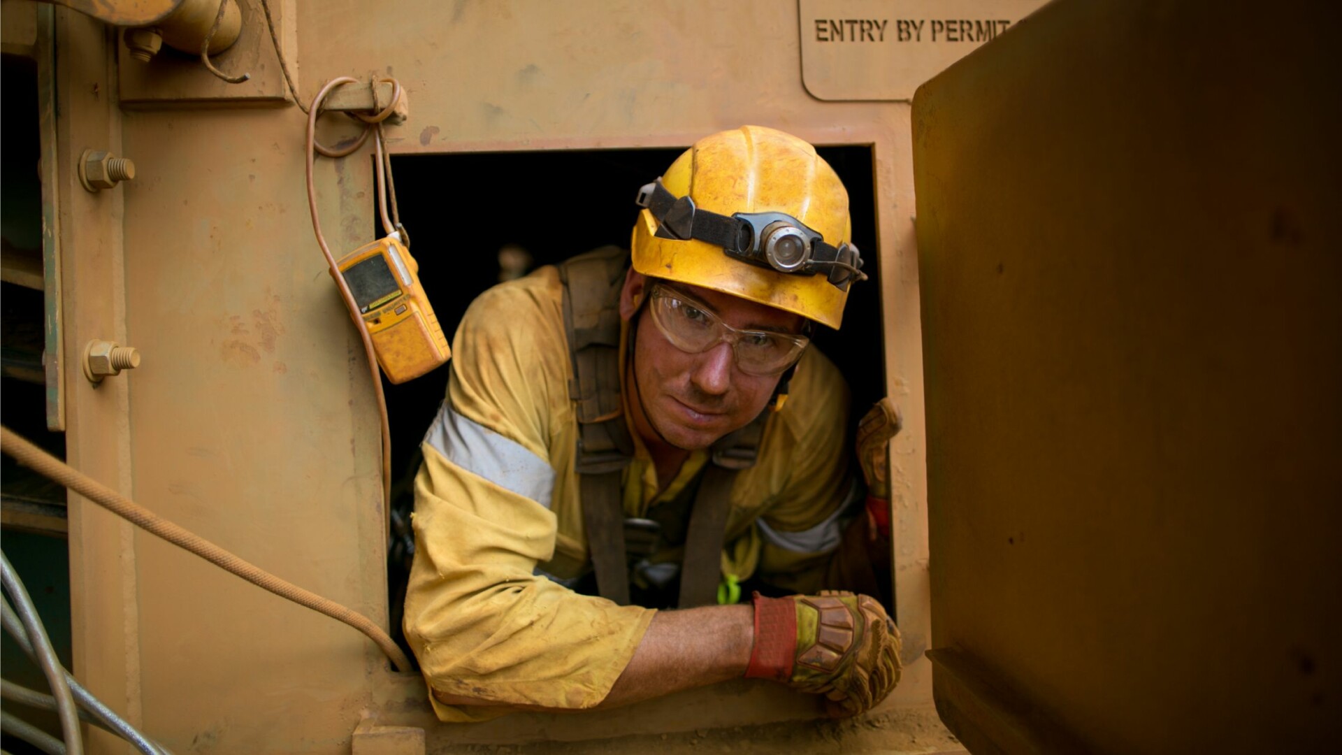 A worker wearing a helmet and other PPE leans out of the doorway of a confined space. A gas detector hangs near the doorway.