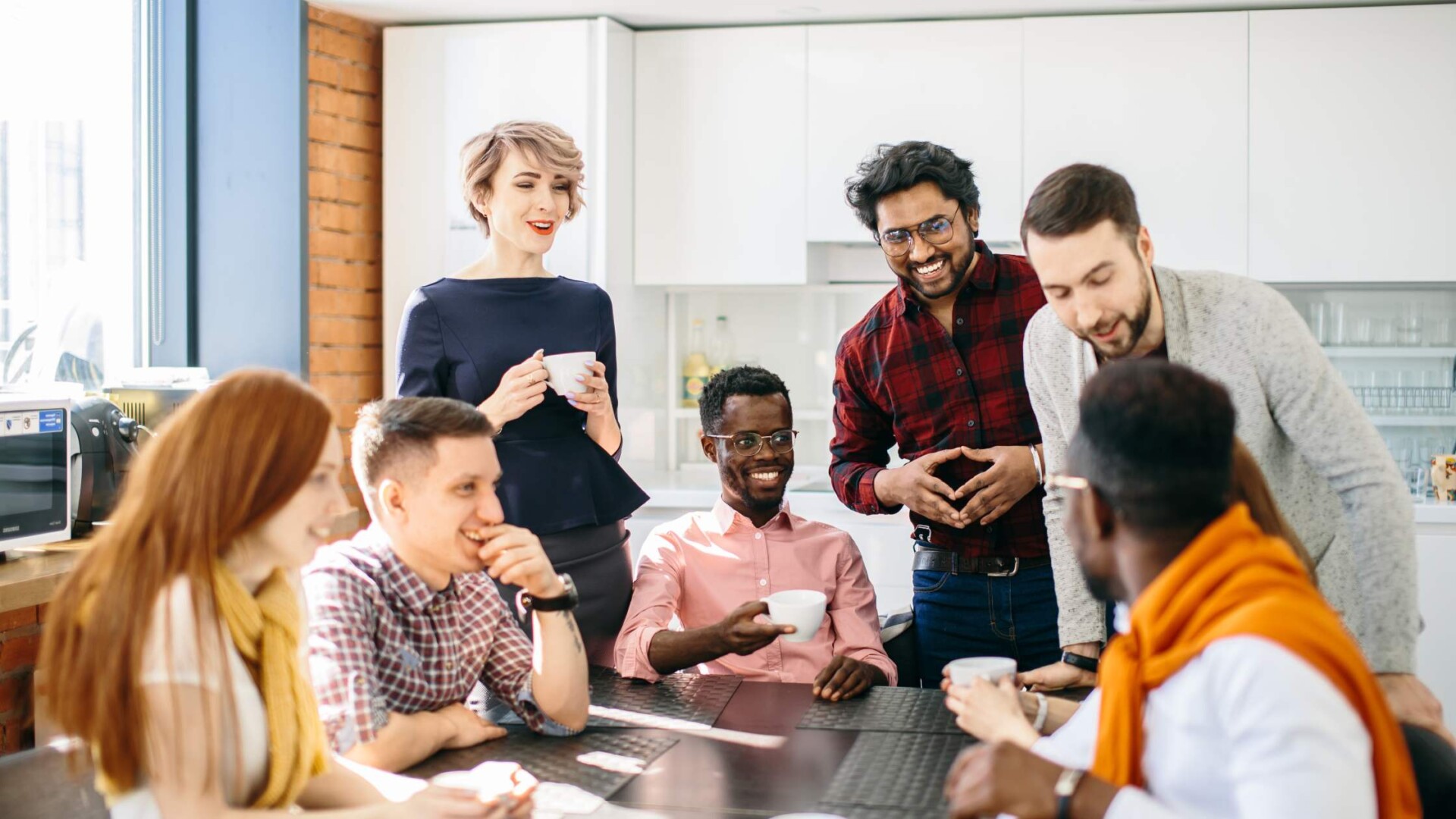 A group of several coworkers sitting and standing around an office kitchen table socializing and holding coffee mugs.