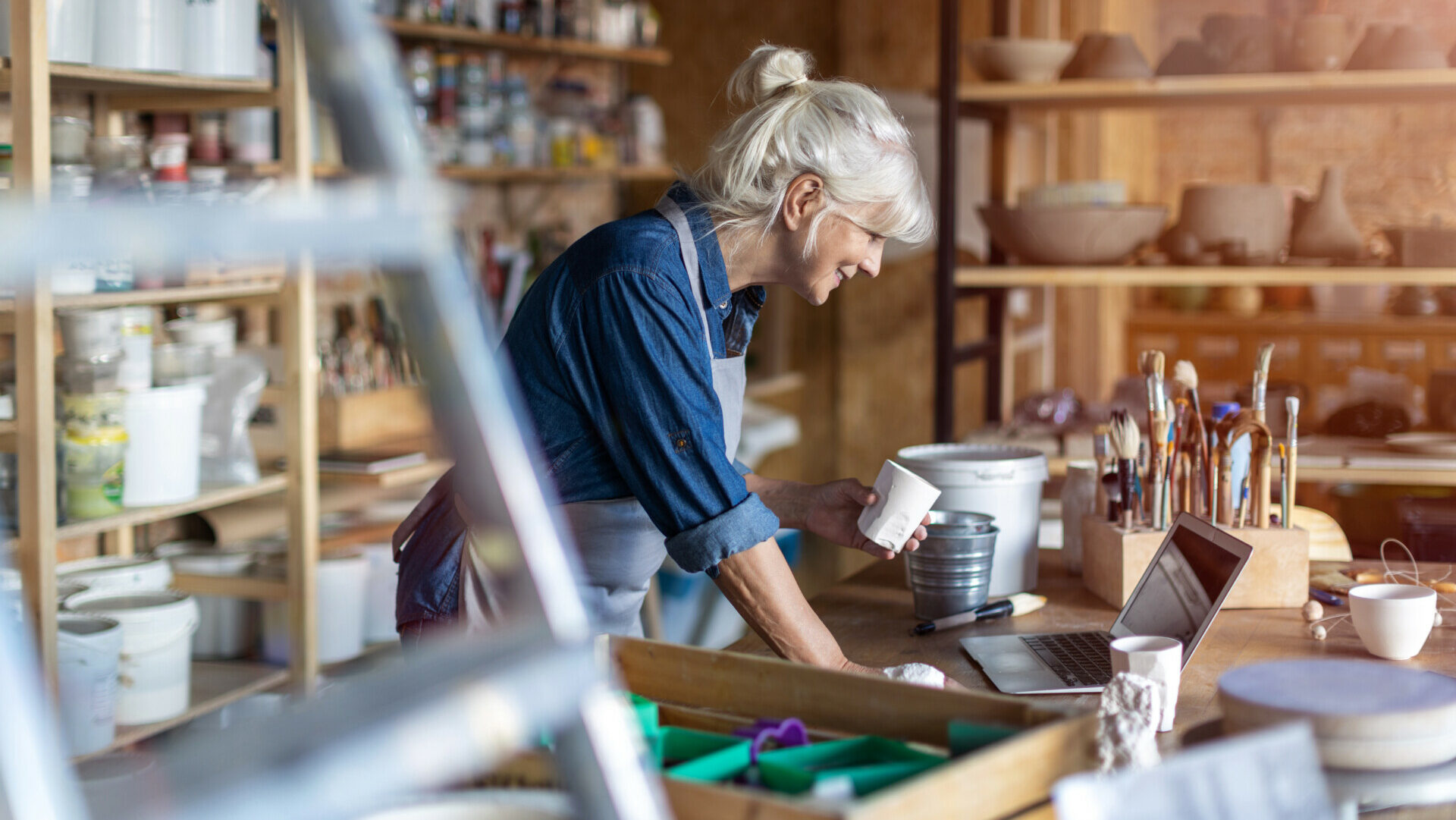 An older woman standing in her pottery studio, holding an unfinished piece and smiling as she looks at her work laptop.