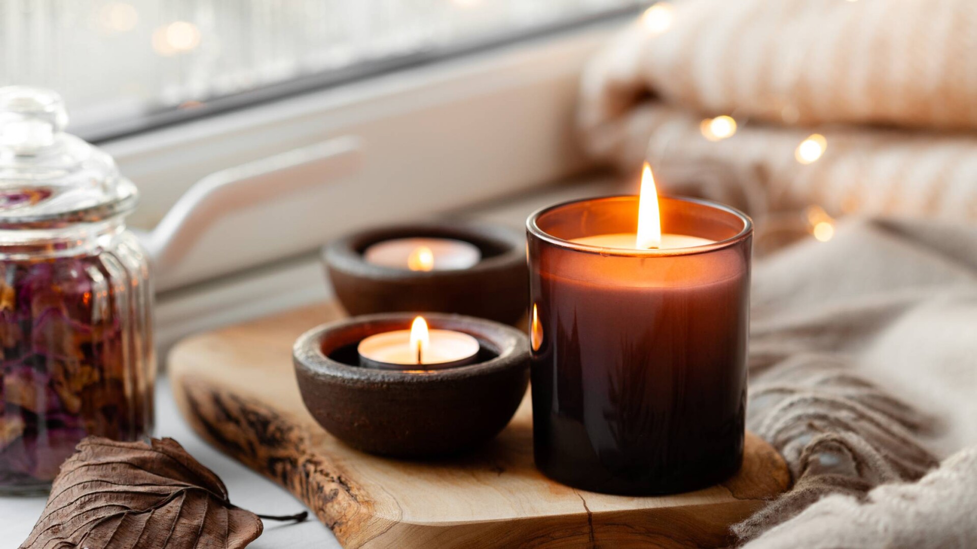 A cozy arrangement of three lit candles by a windowsill surrounded by plush blankets and unlit candles.