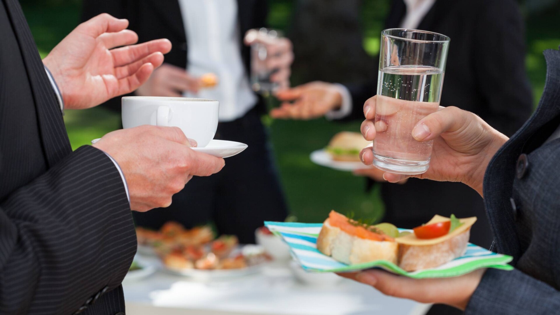 Two business people are holding drinks and small snacks at an outdoor event. They are talking near the food table.