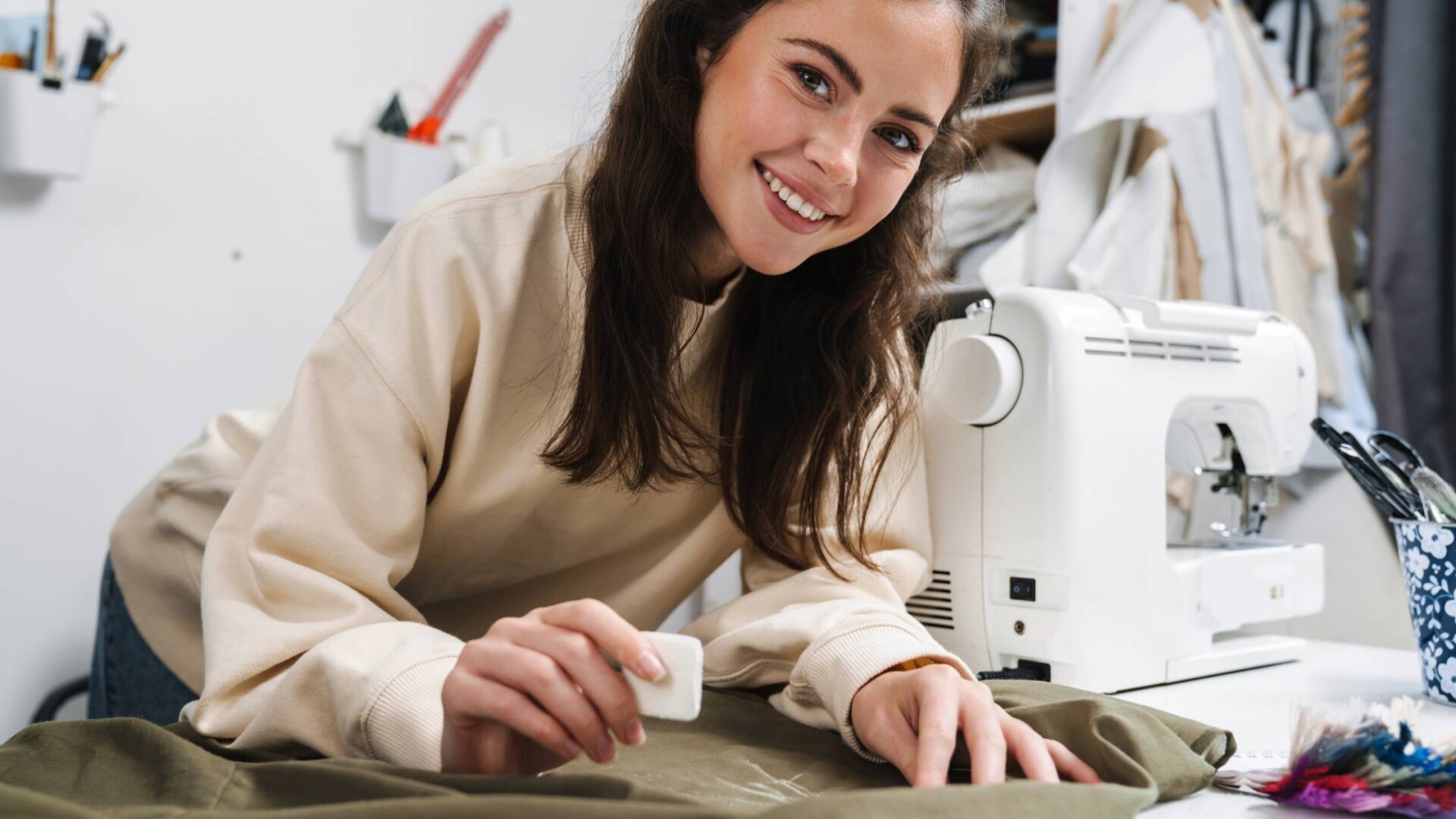 A young woman smiling as she leans over a sewing project and marks the cloth with fabric chalk next to her sewing machine.