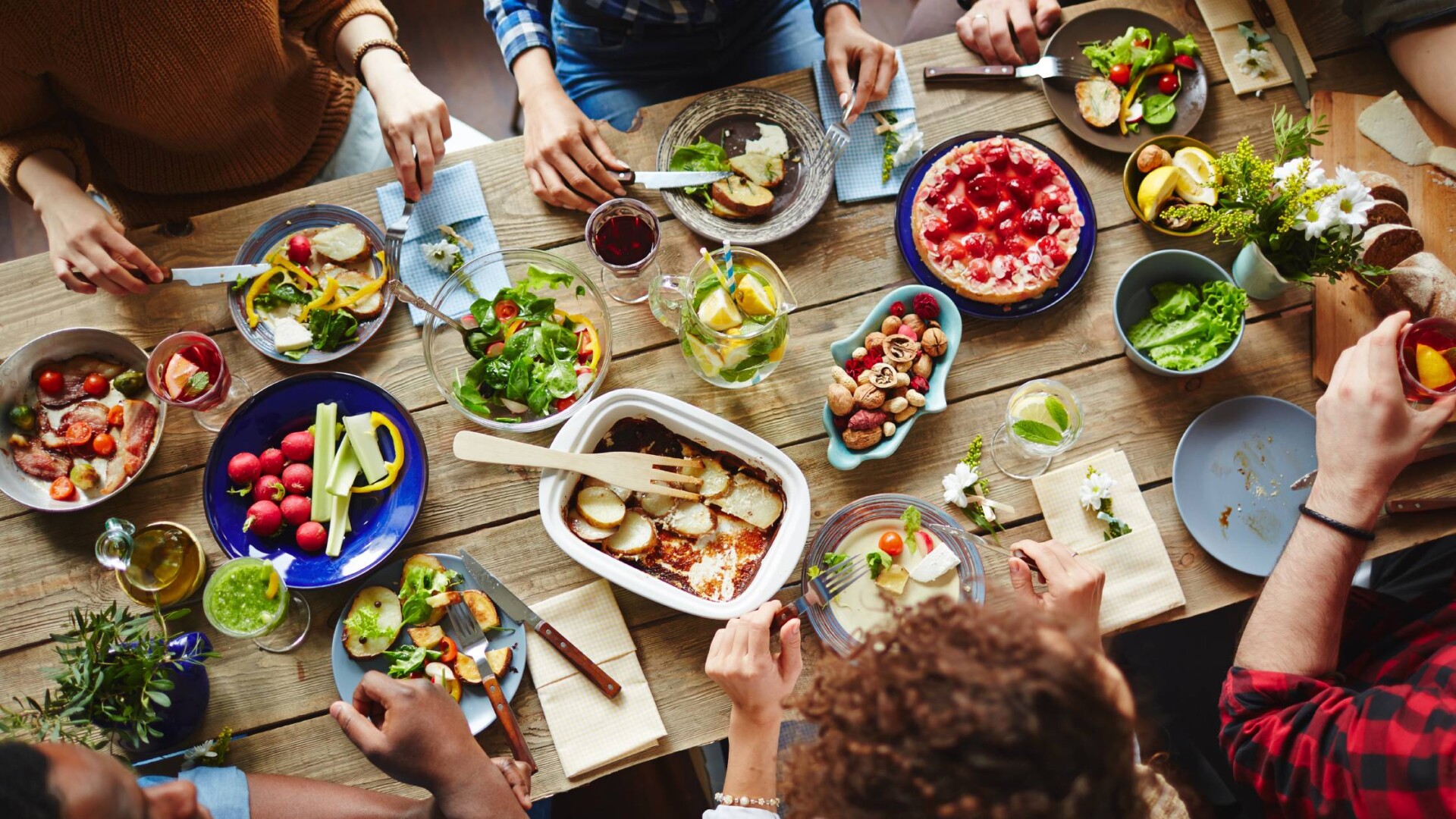 A top-down view of six people eating a meal together at a rectangular table. The dishes include brightly colored vegetables.