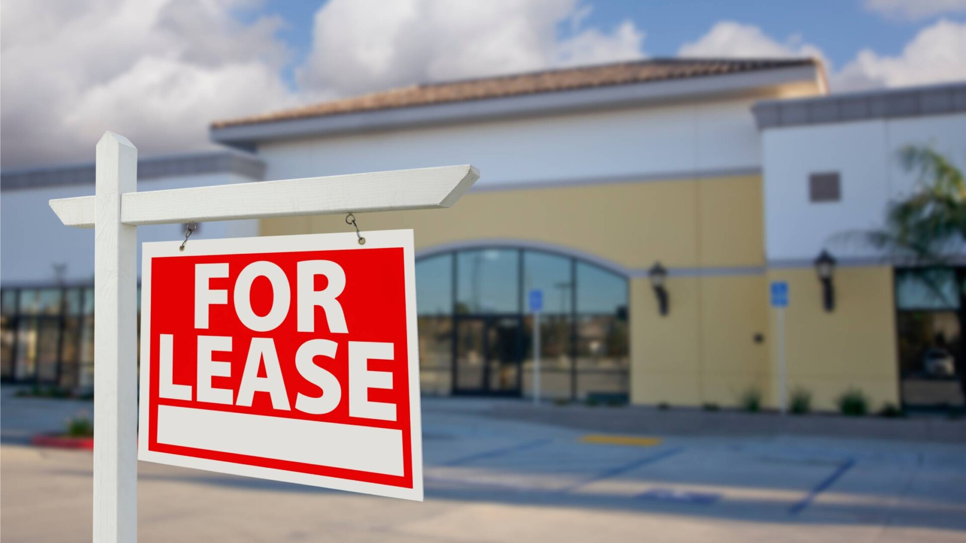 A For Lease sign is in front of a commercial building and an empty parking lot. The building has dark storefront windows.