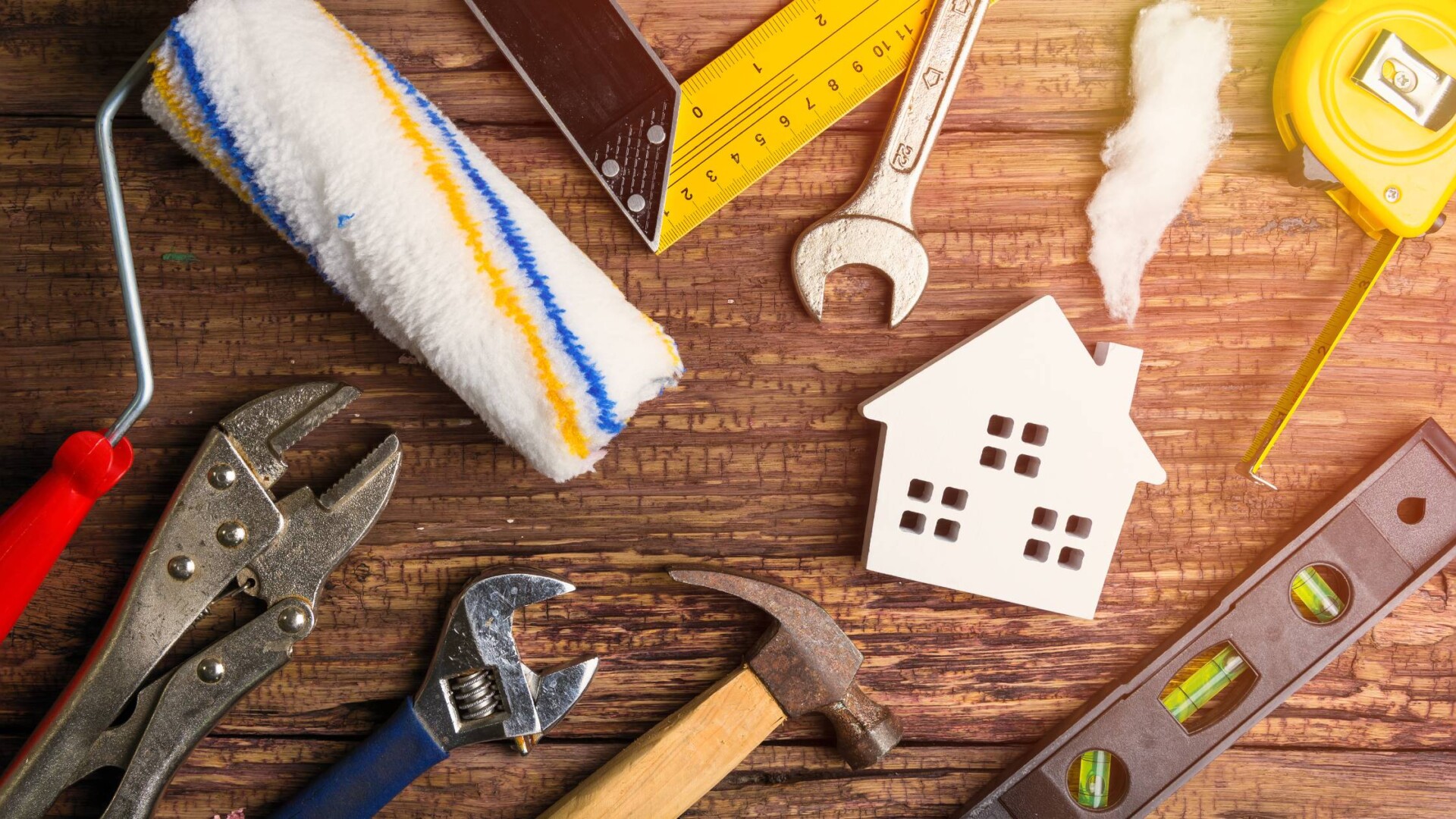 A white wooden cutout of a home surrounded by tools, including a paint roller, wrench, hammer, level, and measuring tape.