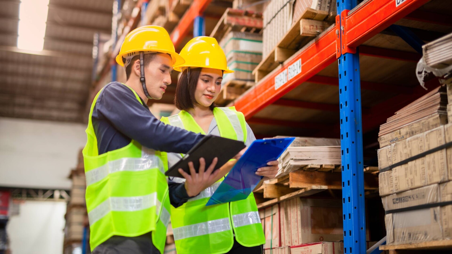 A man and woman wearing yellow hard hats and high visibility vests look at clipboards inside a warehouse.