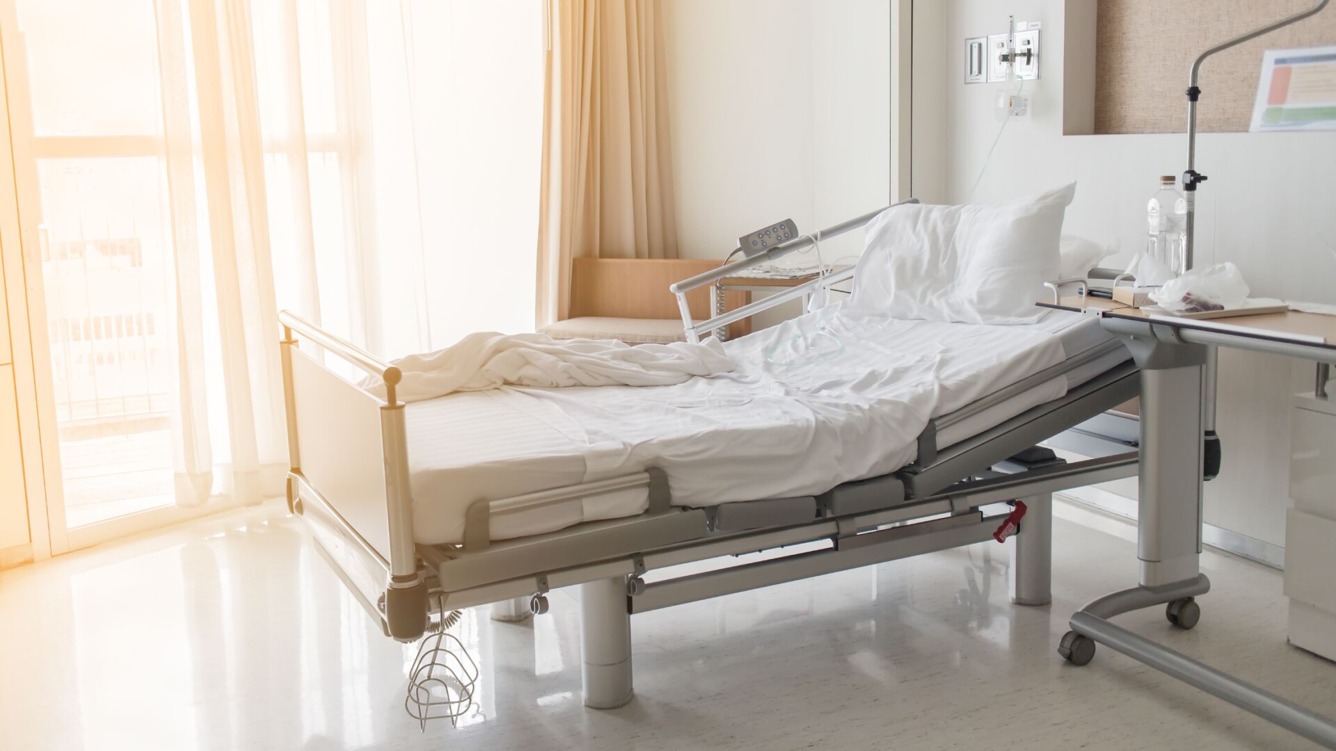 A white and grey adjustable hospital bed has wrinkled white sheets and sits in a room. The curtains are open.