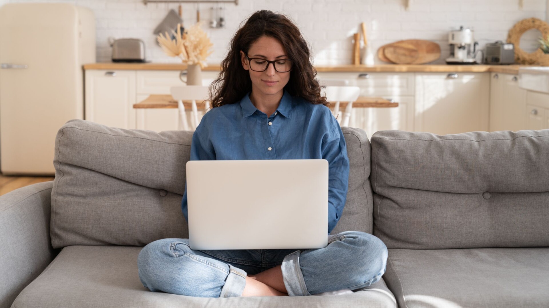 A woman wearing a denim shirt, jeans, and black frames sits on a gray couch, working on a silver laptop.