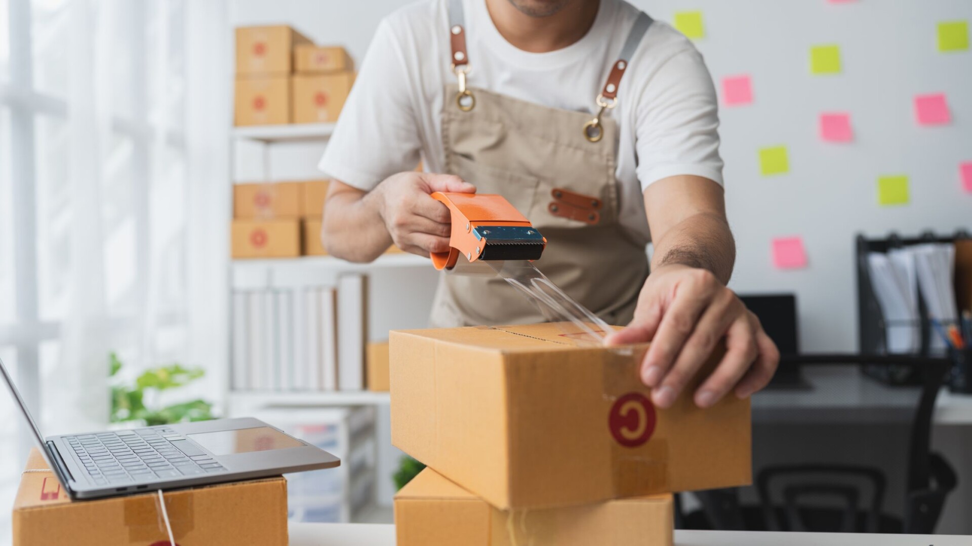 A business worker using a tape gun to securely seal a brown cardboard box with a red logo on its side.