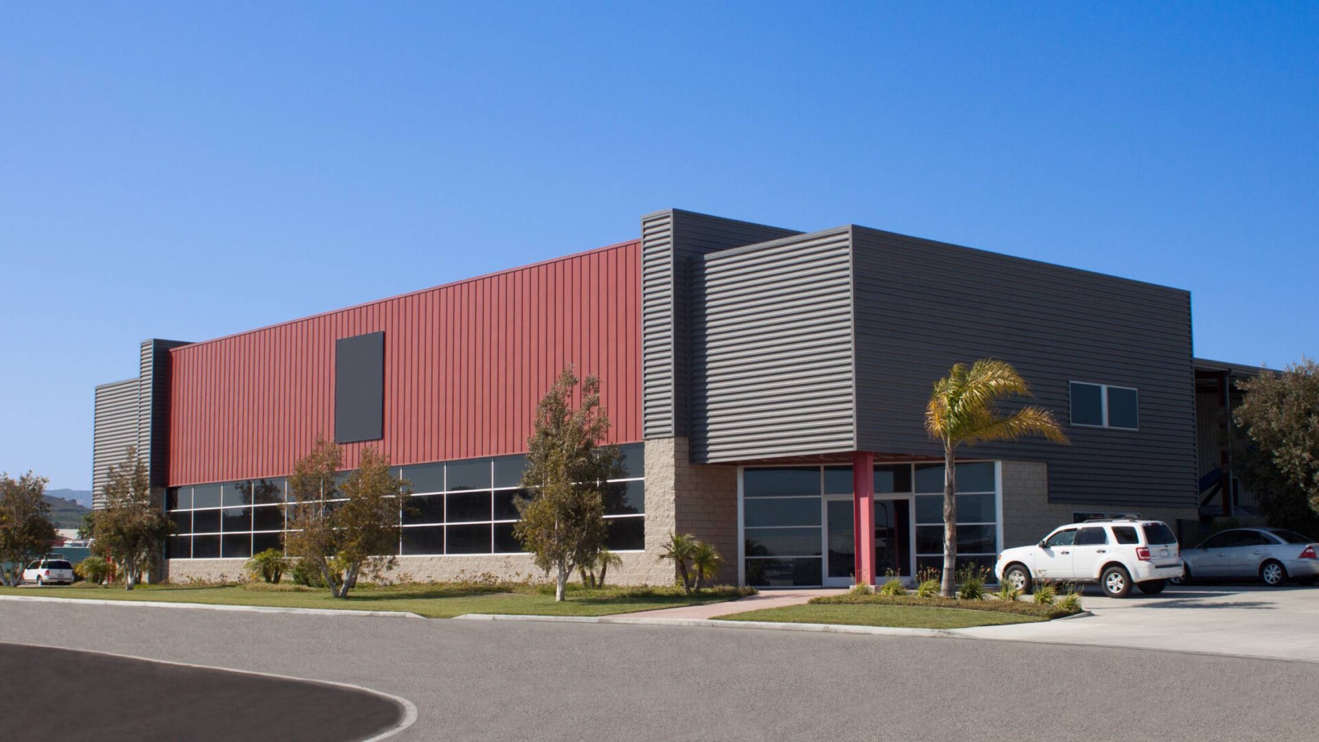 A large commercial business facility with red and brown siding. There are two cars parked in the front of the building.