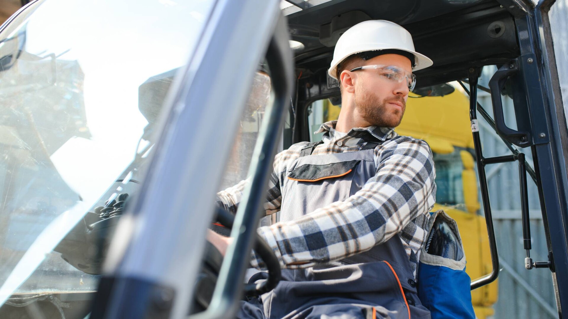 A man wearing a white hard hat driving a forklift. He is looking toward his left as he turns the steering wheel.