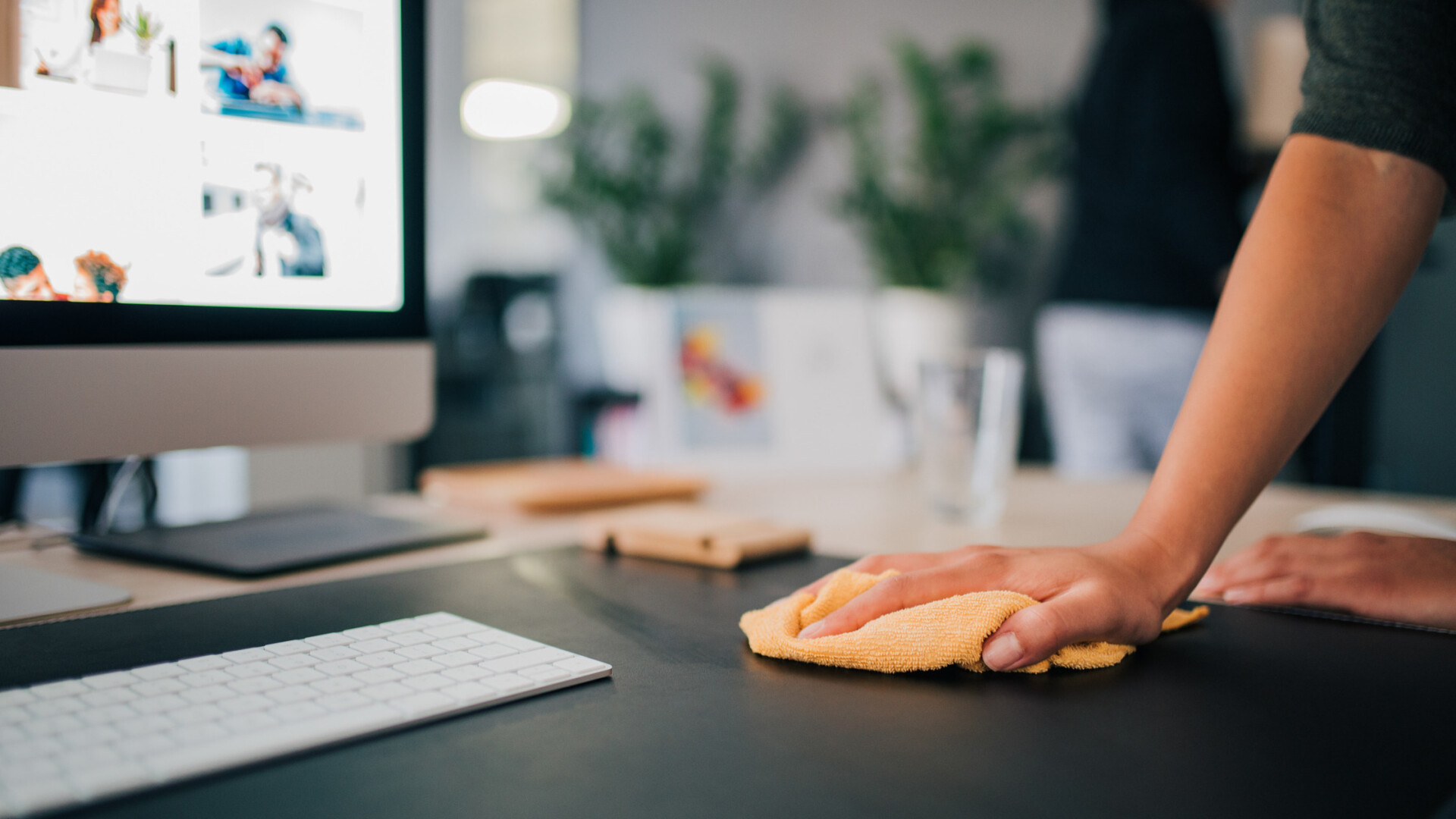 A person uses an orange rag to wipe a black desk clean. A computer monitor and keyboard sit on the desk.
