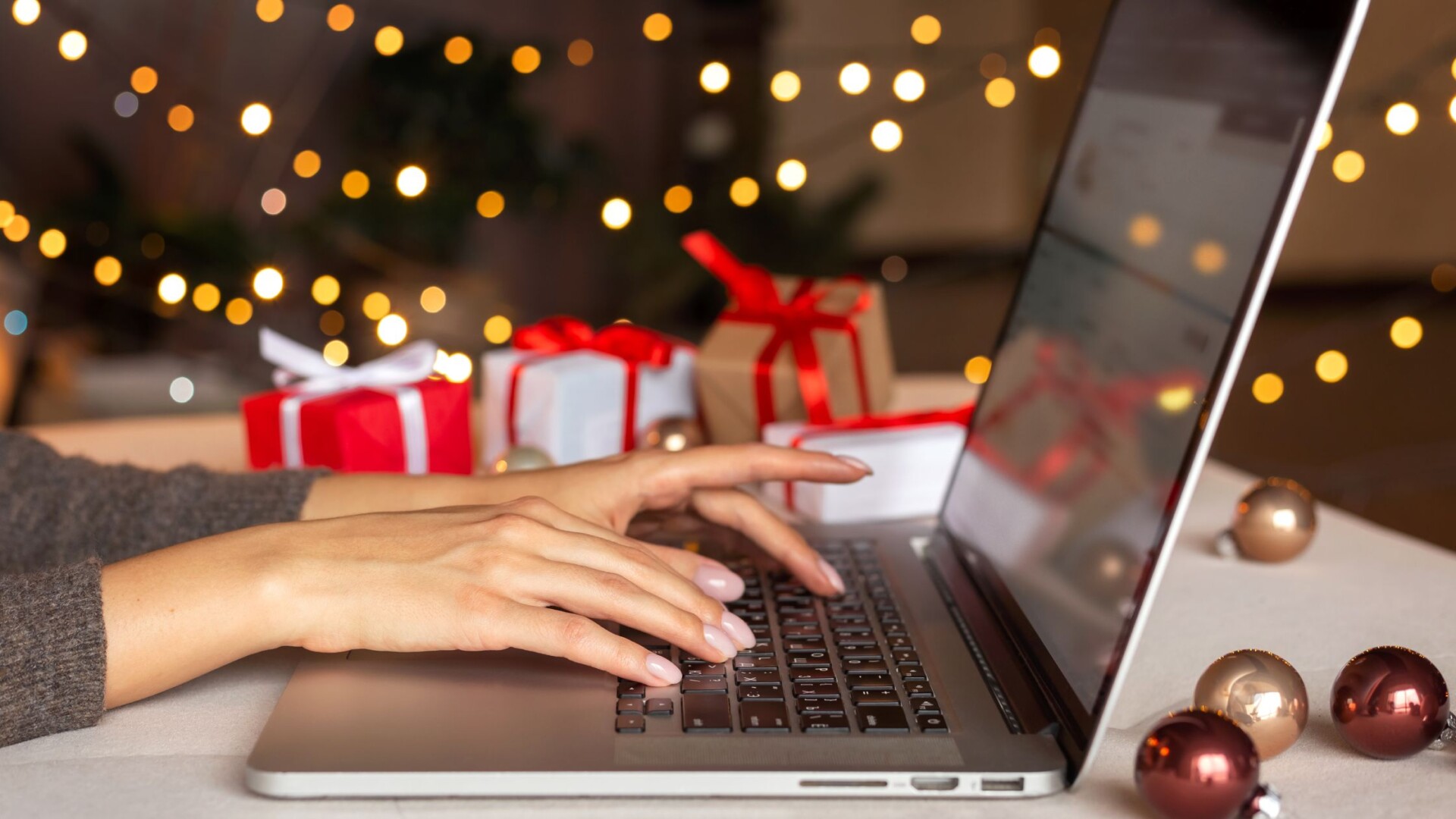 An online shopper sitting at their laptop with small gifts next to them. There are Christmas lights behind them.