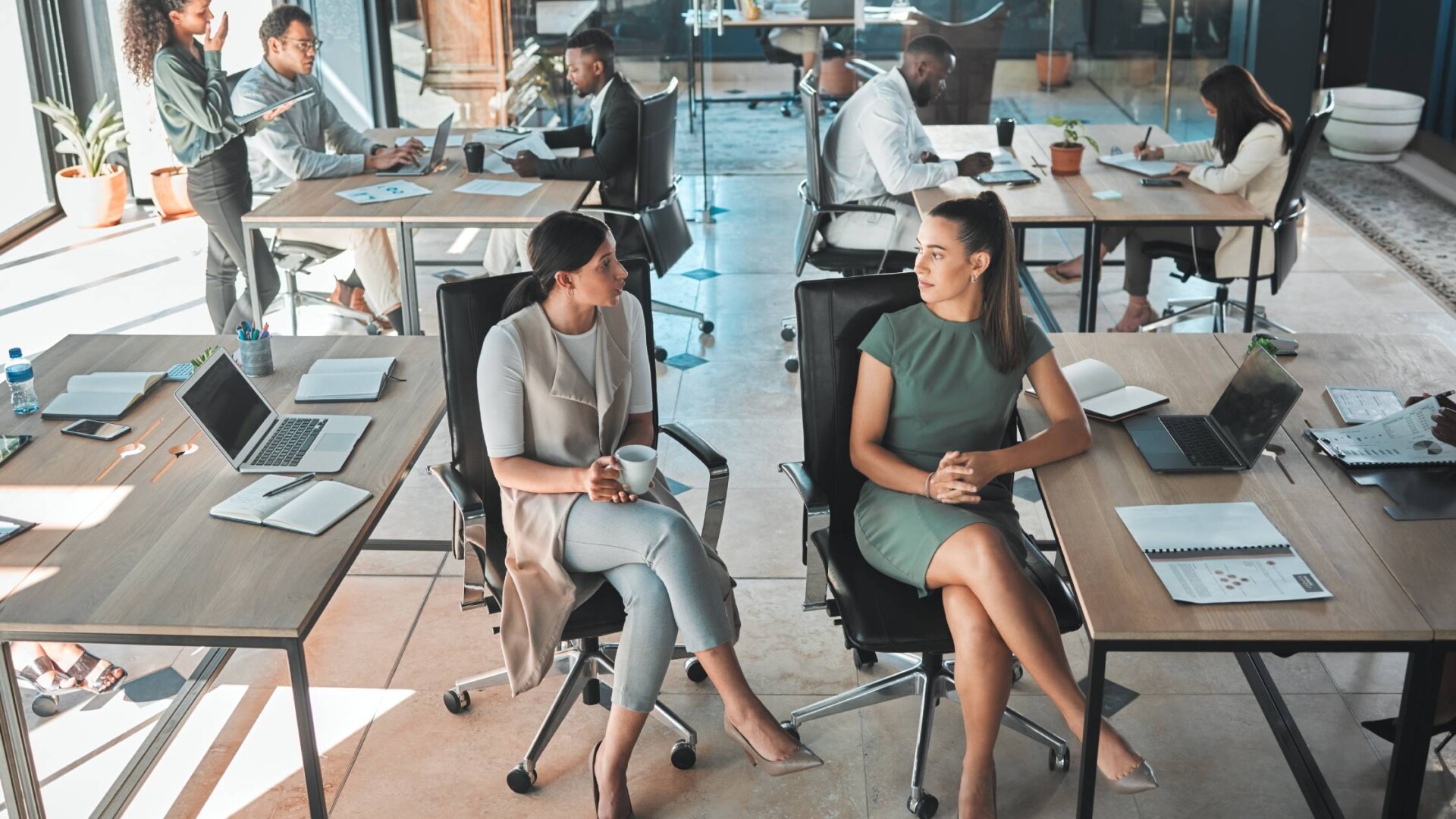 A large, open office space with employees sitting in desk chairs at tables covered with pens, papers, and laptops.