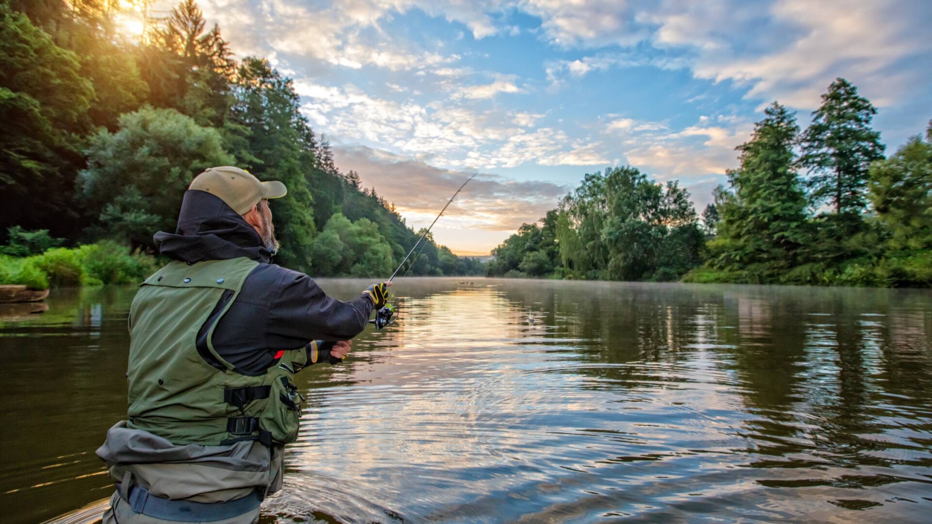 An angler with a green vest who is holding a fishing pole while fishing in the middle of a lake surrounded by trees.