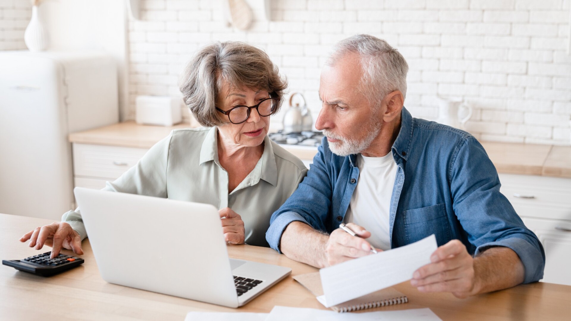 A retired couple sitting at the kitchen table together as they review tax documents. They also have a laptop.