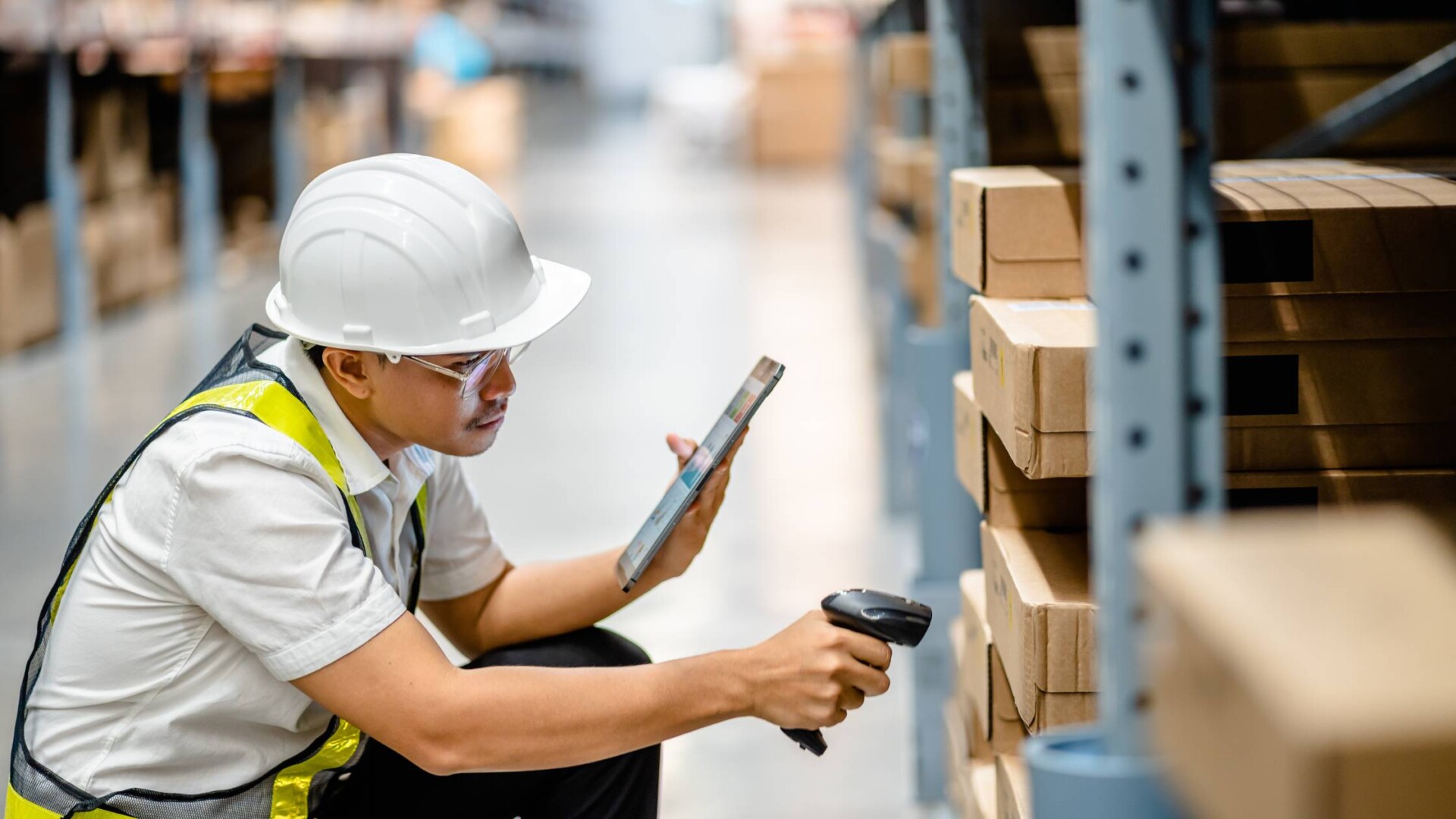 A warehouse employee wearing a helmet and vest as he scans inventory and pallets. He's also using a tablet.