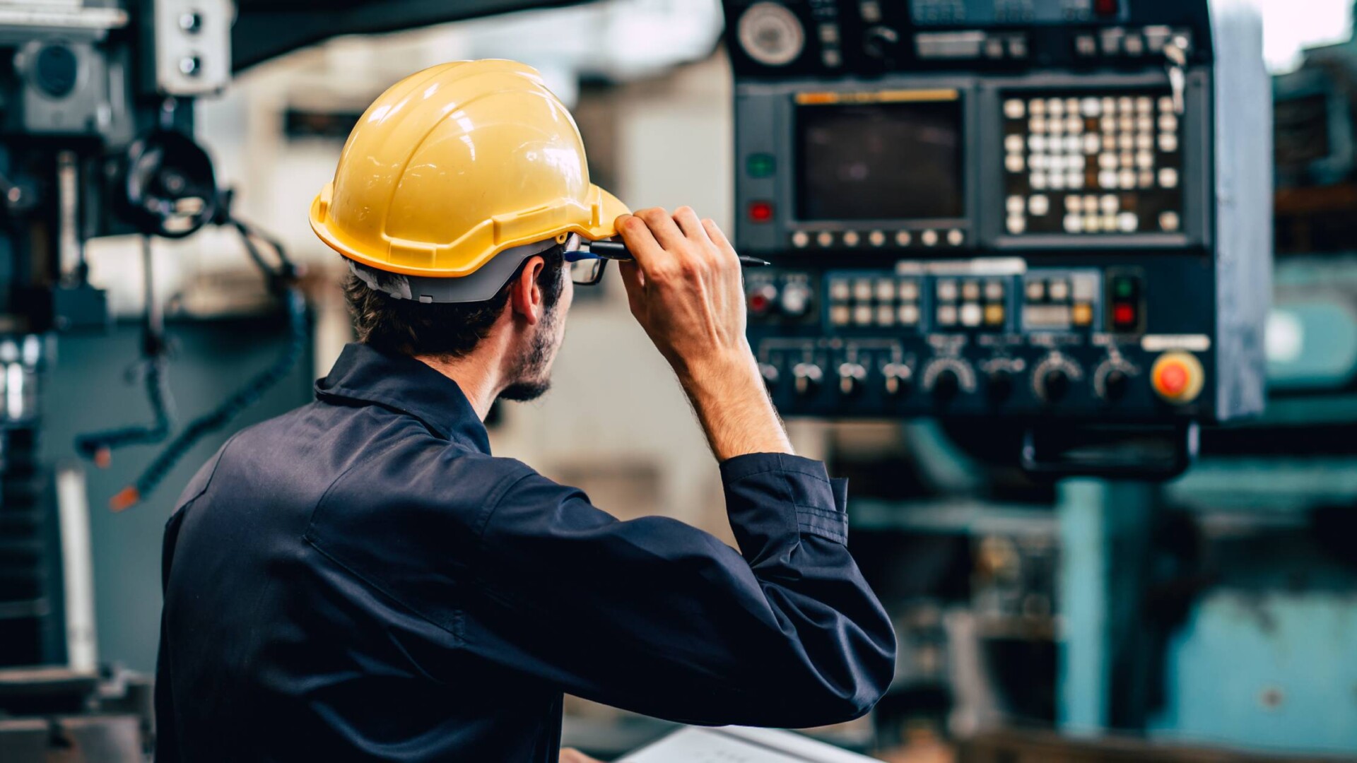A production facility supervisor wearing a yellow safety helmet as he stands in front of a machine's control panel.