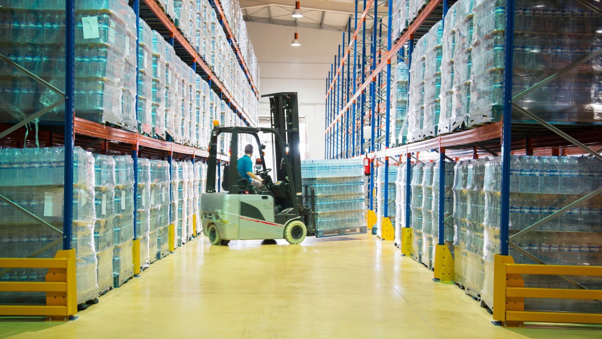 A worker moving pallets with a forklift in a warehouse. They're carrying multiple cases of large plastic water bottles.