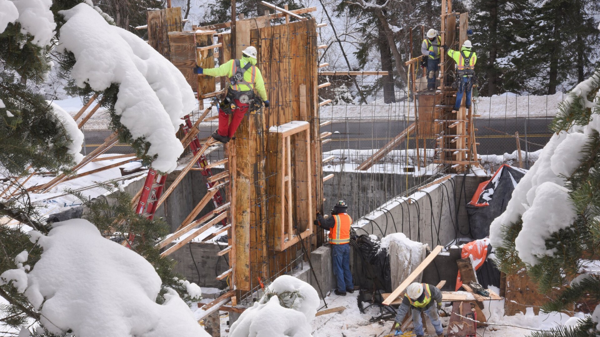 Construction workers build a structure outdoors in a snowy environment. Heavy snow rests on evergreen trees in the foreground.