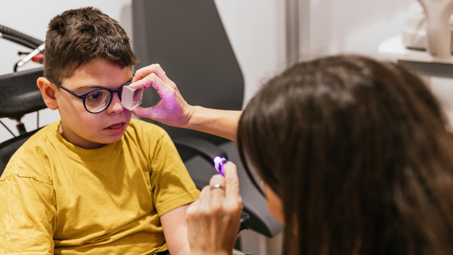 An optometrist uses a purple light to check a child's eyes in the exam room. The child sits in a wheelchair.