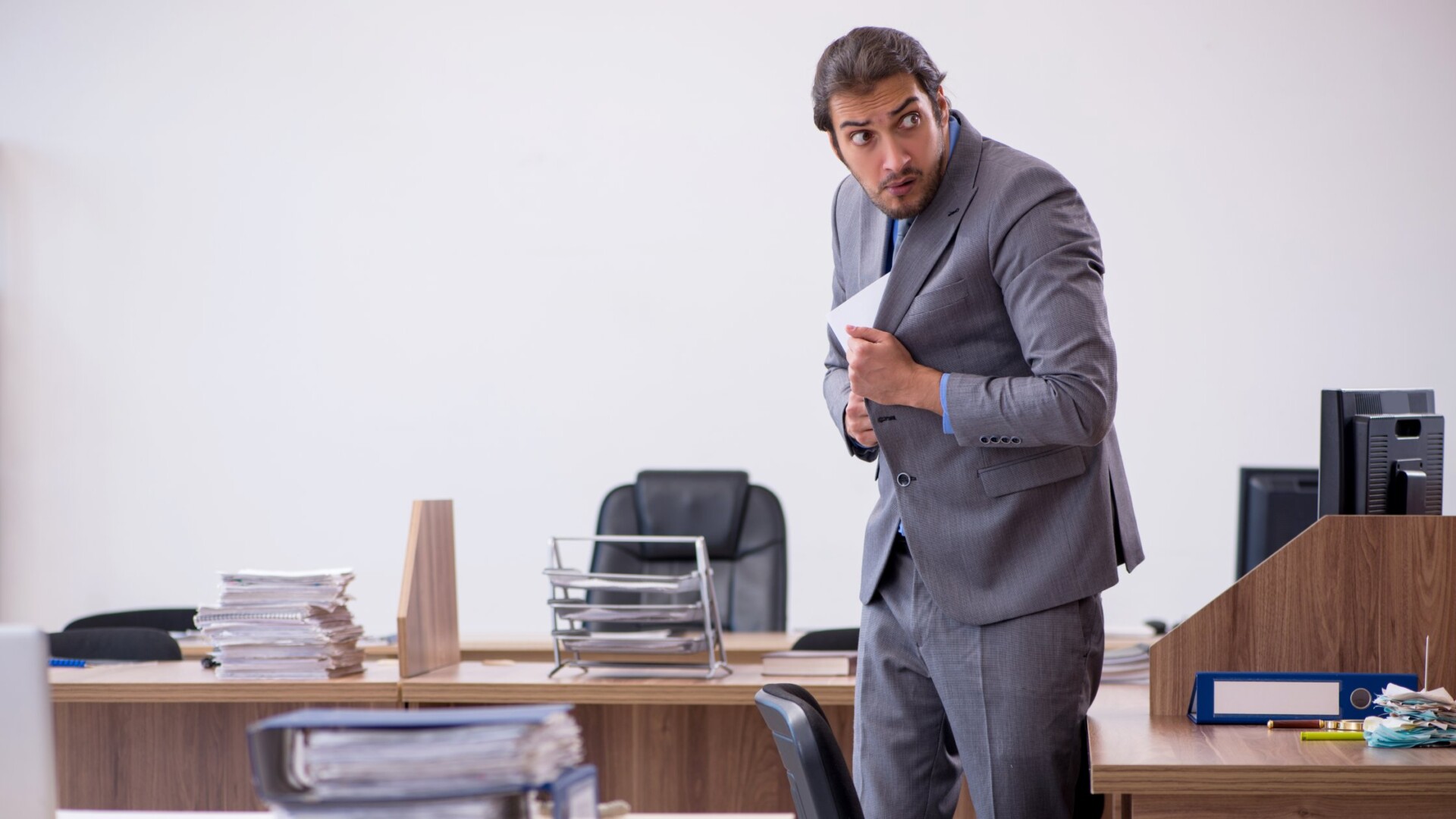 An office employee hides documents in his suit jacket. He's standing near desks with documents and binders.