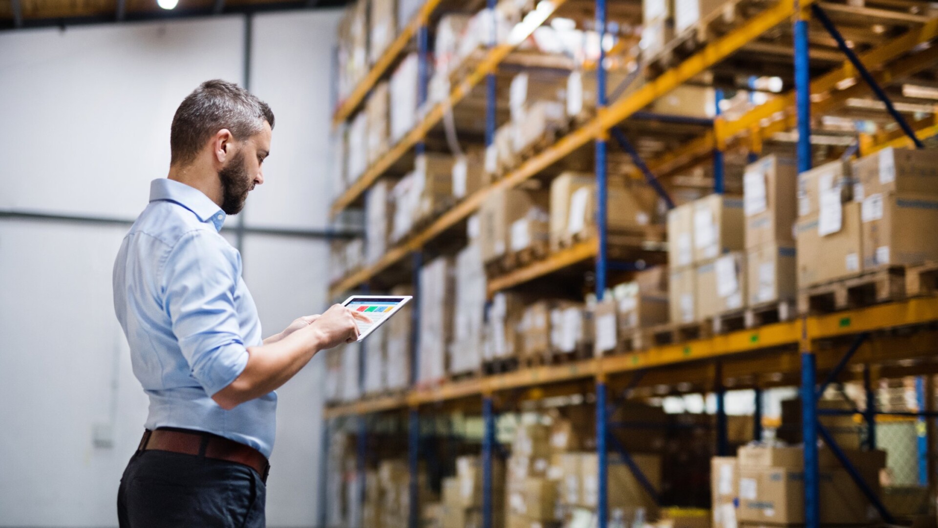 A man in business attire is using a tablet while standing in front of warehouse racks full of boxes.
