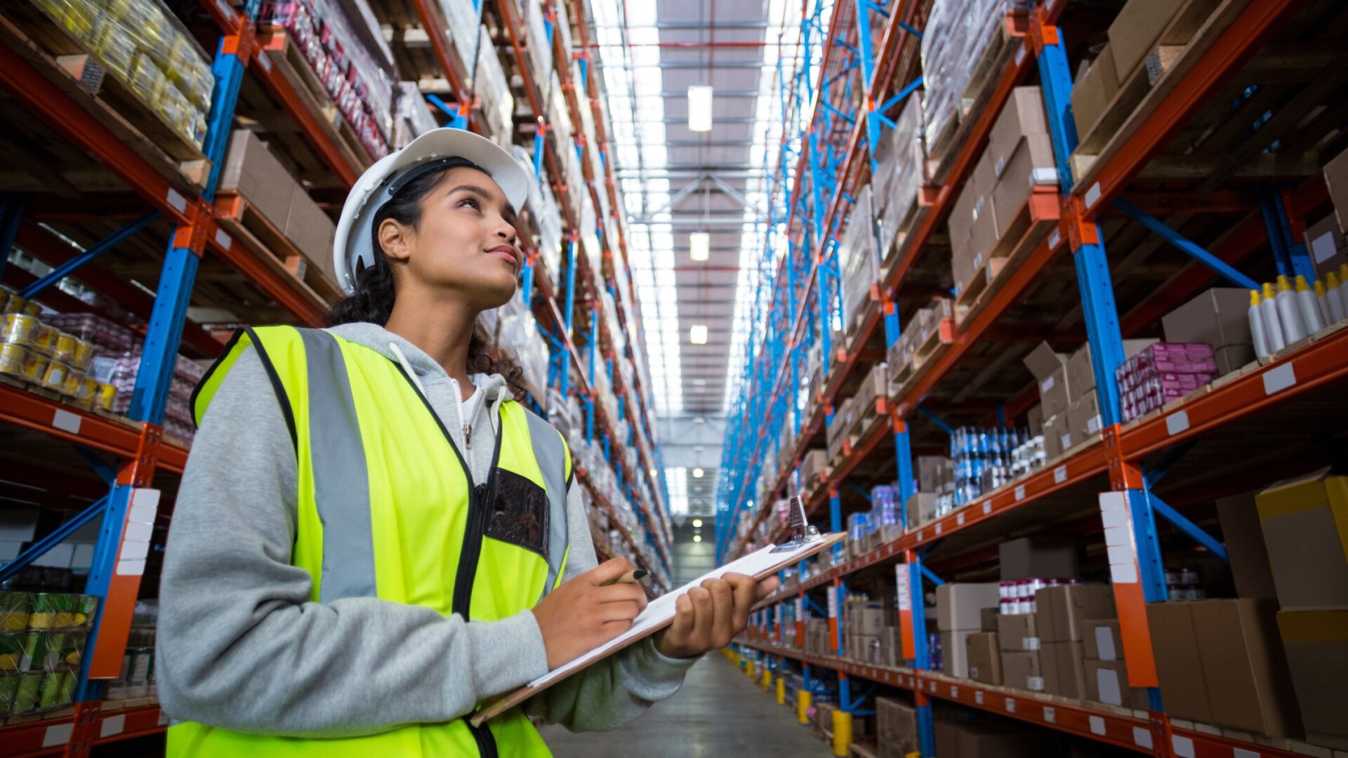 A woman in a white hard hat and yellow vest holds a clipboard and looks up at products shelved in a warehouse.