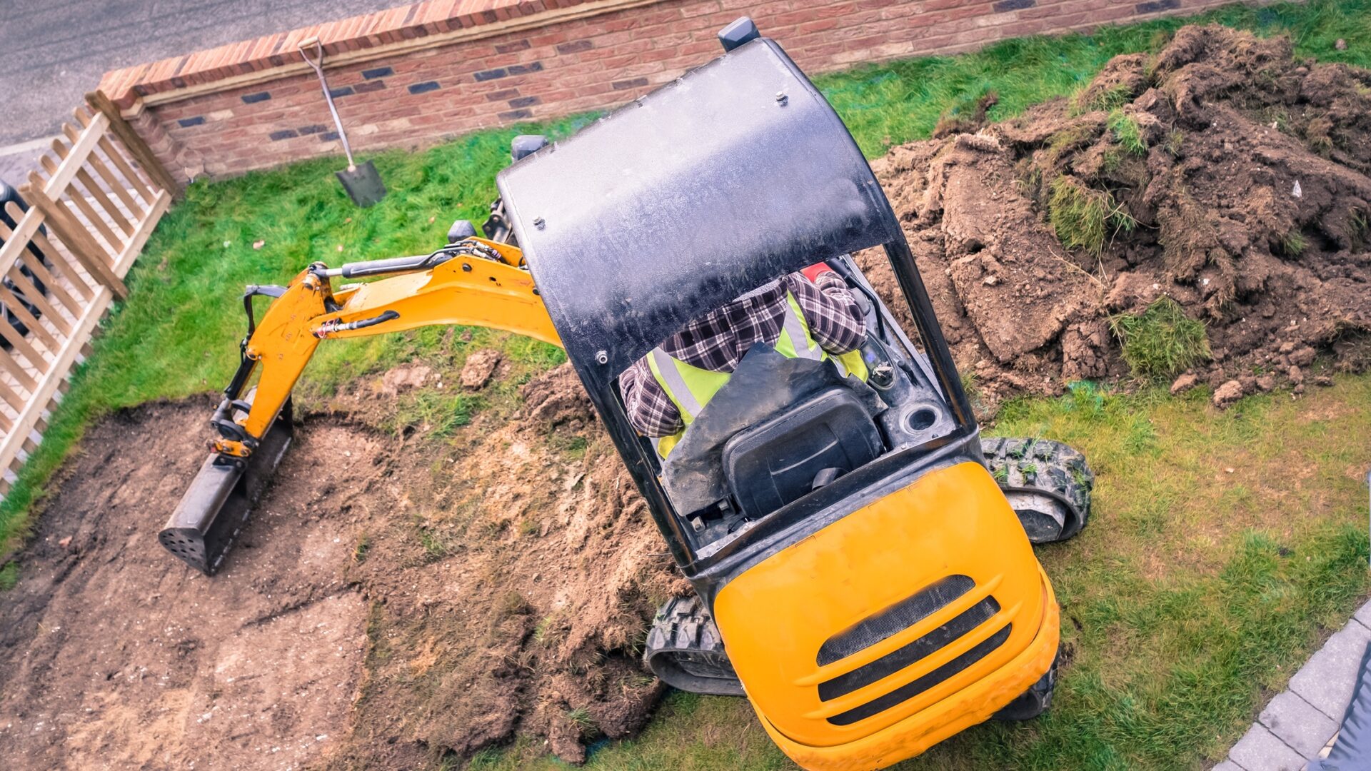 A worker removes turf in a small grassy area with a mini excavator. A pile of dirt and turf is next to the machine.