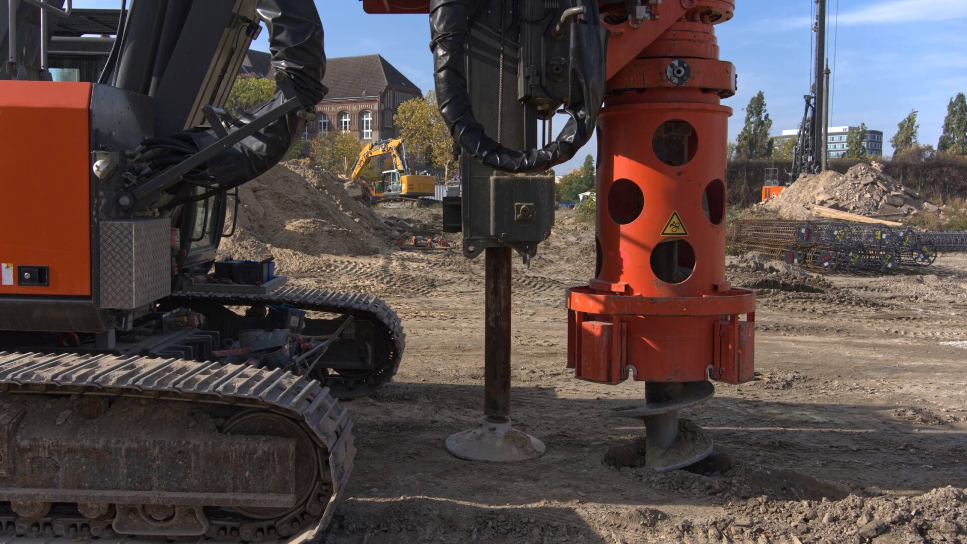 A large drilling machine with an orange auger attachment at a construction site with dirt piles and other equipment.