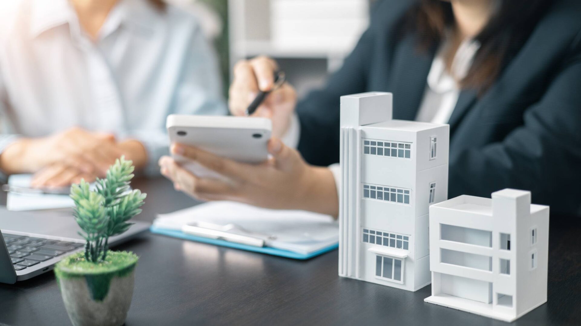 Two people sitting at a desk with models of buildings, a small potted plant, a laptop, and a clipboard with documents.