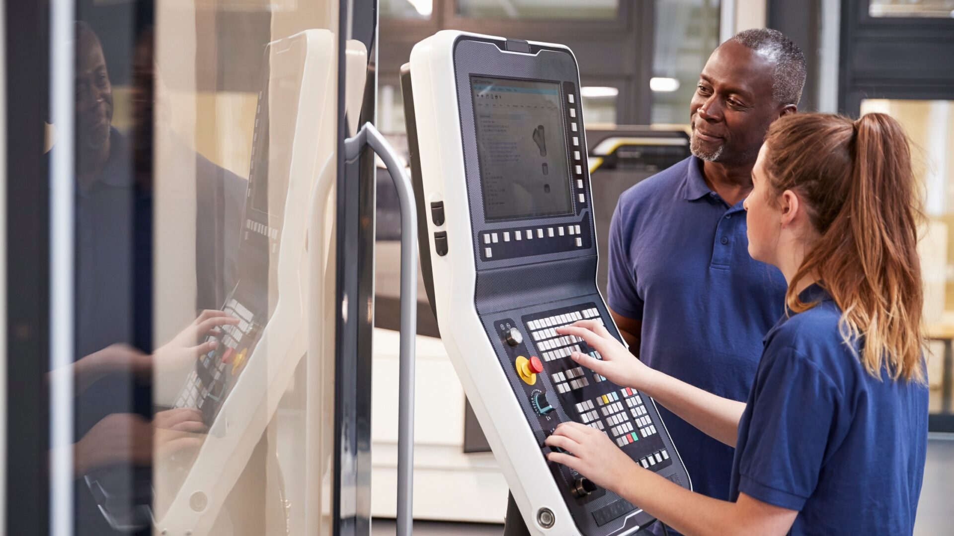 Two CNC machine operators standing at the machine's computer to work out a problem. They're in their work uniform.