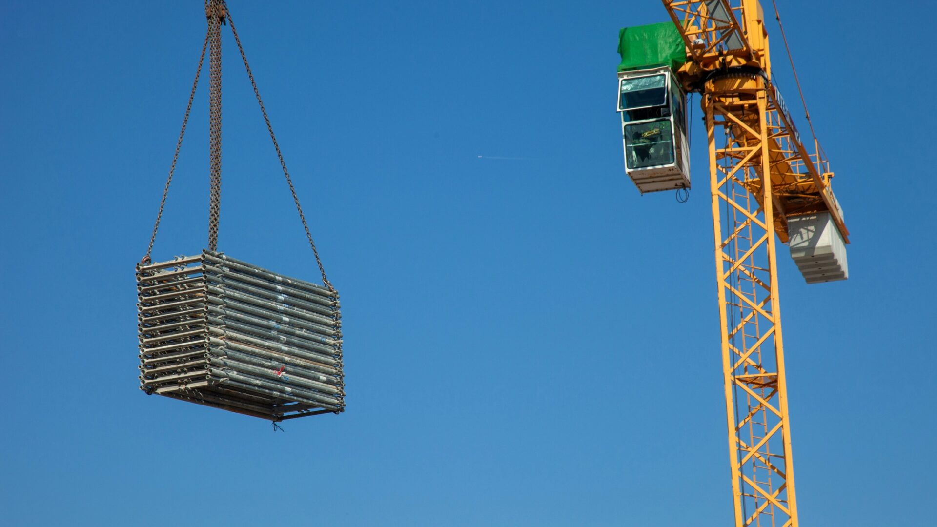 A large yellow construction crane with a clear blue sky behind it. The crane is lifting several metal pieces rigged together.