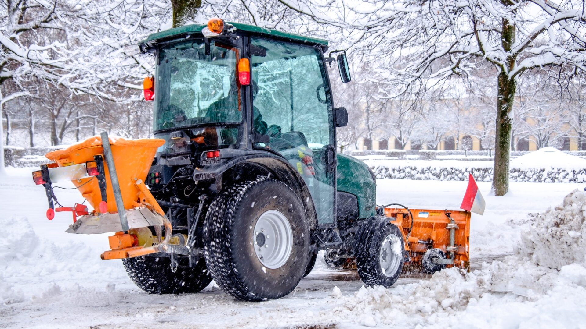 A green compact tractor using a front-mounted snow plow attachment to clear a safe walkway in the snow.