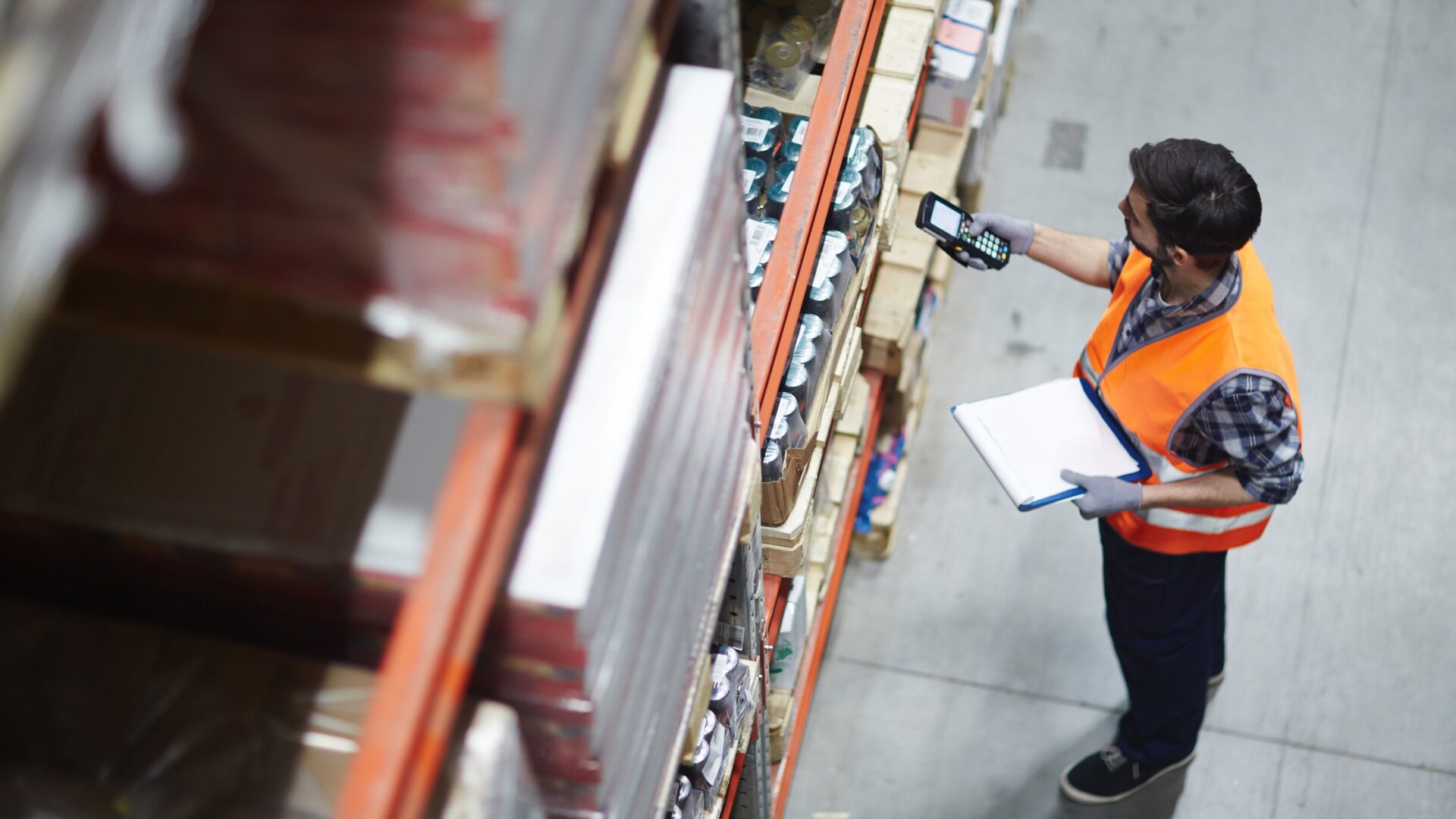A warehouse worker in a safety vest holding a clipboard and using a handheld scanner to check inventory on the shelves.