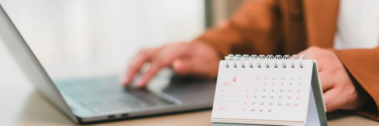 A close-up of a person in a brown jacket using a laptop at a desk with a small calendar sitting on it.