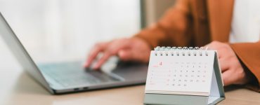 A close-up of a person in a brown jacket using a laptop at a desk with a small calendar sitting on it.