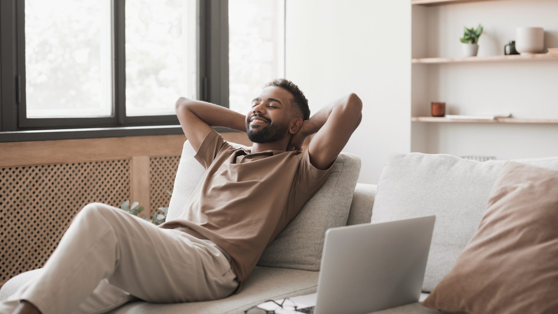 A man smiling and relaxing on a sofa with his foot on a table. His work computer and glasses rest beside him.