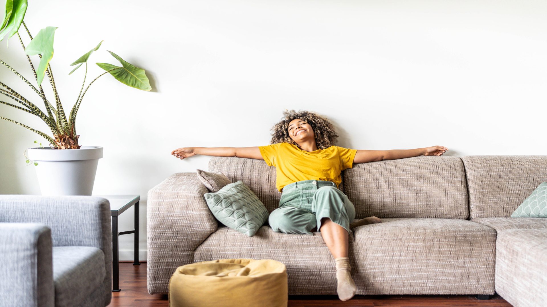 A woman sits and relaxes on a couch in her home. A tall plant stands nearby and sits on a glass table.