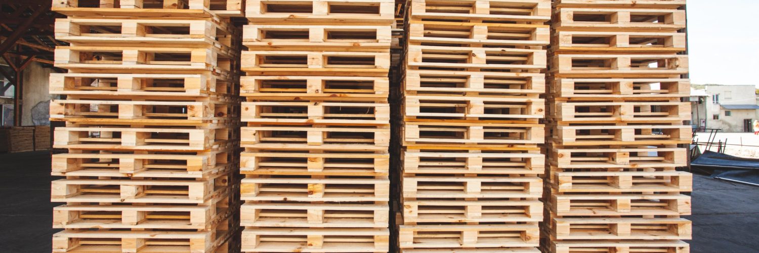 Four stacks of wooden shipping pallets stacked to the side of a logistics facility. The pallets have new wood, ready for use.