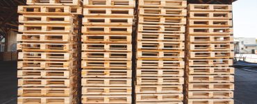 Four stacks of wooden shipping pallets stacked to the side of a logistics facility. The pallets have new wood, ready for use.