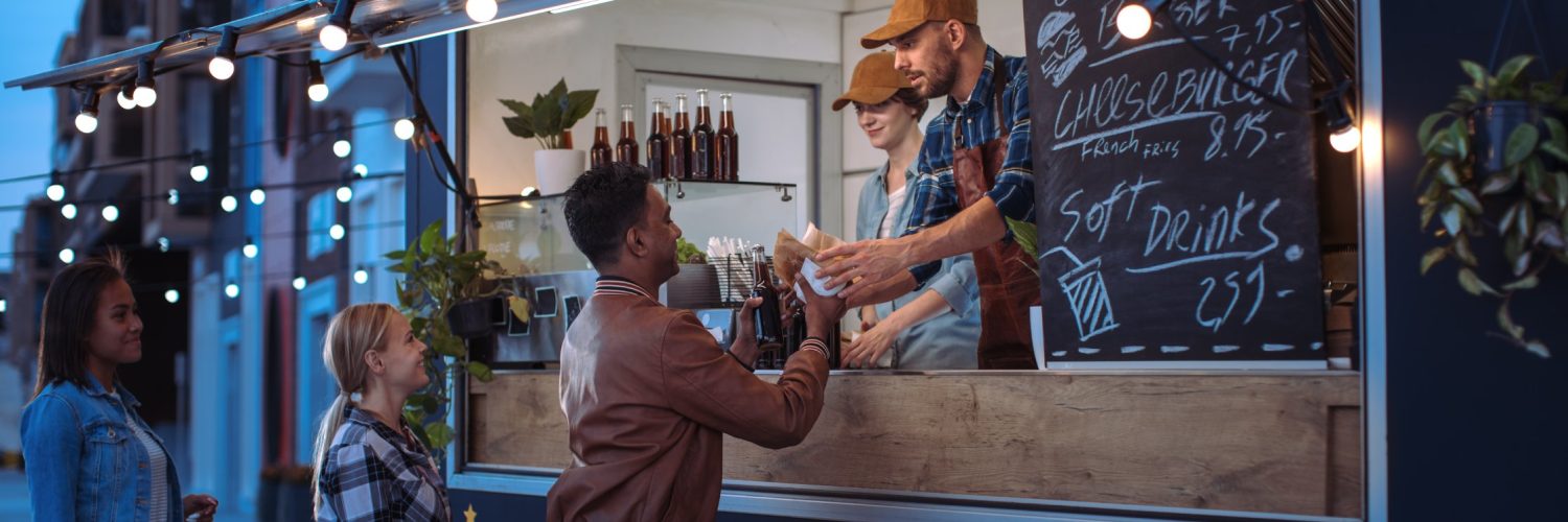 Three people line up outside a black and blue food truck at night. A worker hands food to the person in front.