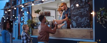 Three people line up outside a black and blue food truck at night. A worker hands food to the person in front.