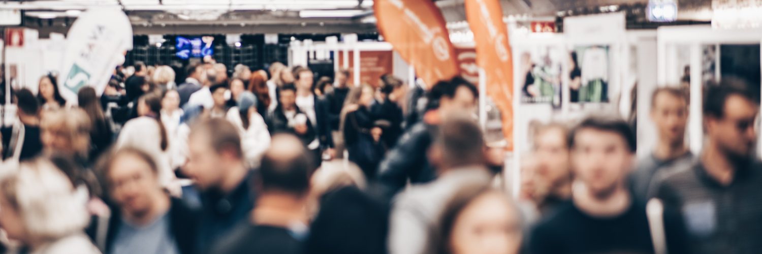 A blurred crowd of trade show attendees walks through an event hall, heading toward various business booths.