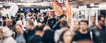 A blurred crowd of trade show attendees walks through an event hall, heading toward various business booths.
